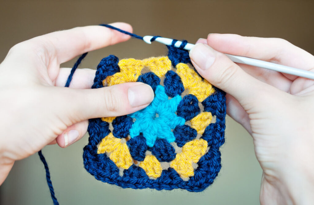 Crocheted square, navy, gold, teal colors. Hands holding crochet hook and yarn.