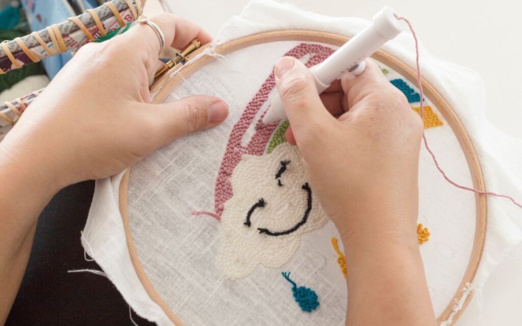 Closeup of hands embroidering a cloud-shaped design on fabric using a hoop and needle tool. Colorful thread used.